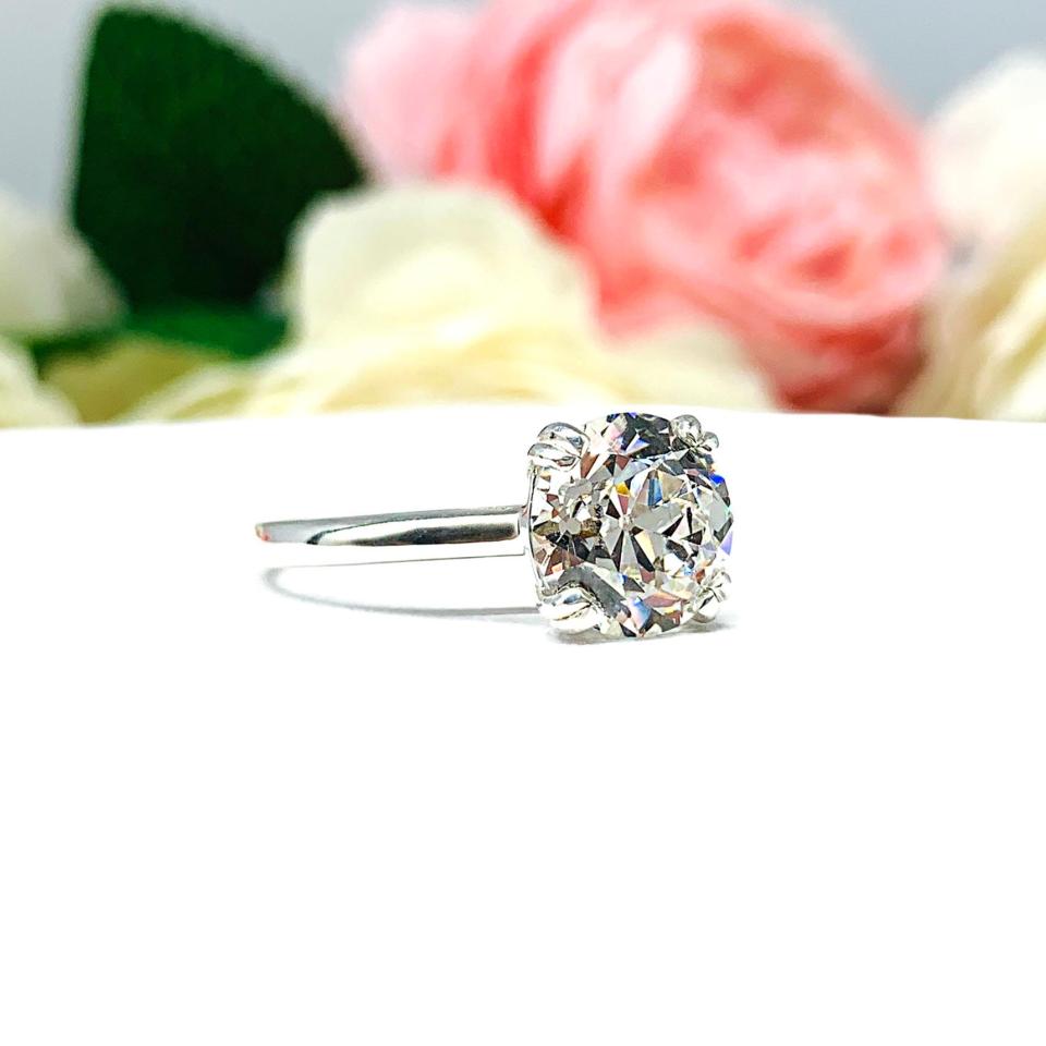 A romantic close-up of a 9mm (3 ct) old European cut near-colorless CZ solitaire engagement ring. Set in a sterling silver split prong cathedral-style mount, the ring gleams with vintage-inspired elegance. Soft pink and white flowers blur in the background, adding a dreamy, bridal ambiance.