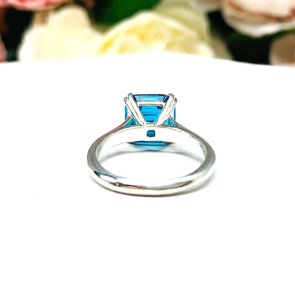 Sterling silver 8mm teal blue asscher cut cubic zirconia solitaire ring. The ring is resting on a white surface with a background of pink and white flowers.