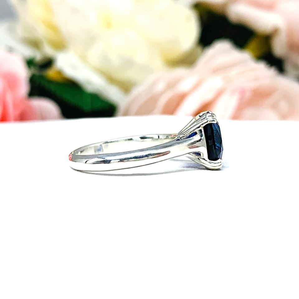 Close-up of a sterling silver cathedral-style ring featuring an 8mm cushion cut lab created blue sapphire center stone.  The ring is facing toward the right to display the polished band.