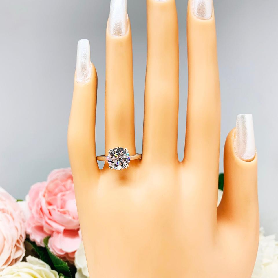 Mannequin hand with long white shimmery nails, displayed against a soft white-gray background with pink and white floral accents. The hand is wearing a colorless round brilliant cut cubic zirconia solitaire ring in a split prong cathedral setting. The ring is made of sterling silver and features a sleek, minimalist design. The setting is simple yet elegant, allowing the center stone to take center stage.