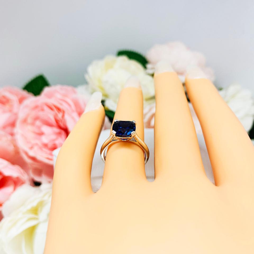 A display hand with long white fingernails laying down and with the top of the hand facing up, displayed against a soft-focus background of pink and white flowers. The display hand is wearing a sterling silver cathedral-style ring featuring an 8mm cushion cut lab created blue sapphire center stone.