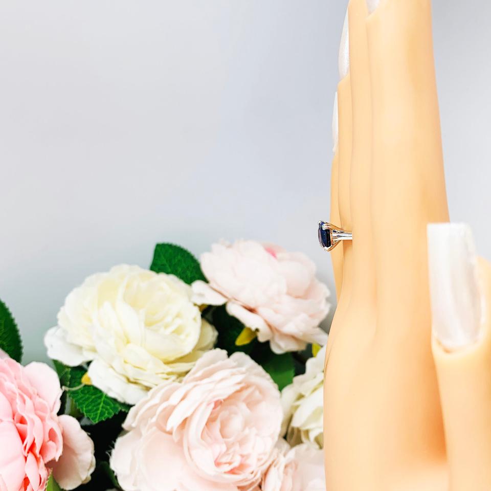 A display hand with long white fingernails standing up and with the top of the hand facing toward the left, displayed against a soft-focus background of pink and white flowers.  The display hand is wearing a sterling silver cathedral-style ring featuring an 8mm cushion cut lab created blue sapphire center stone.