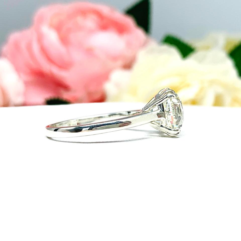 A romantic close-up of a 10mm (4 ct) old European cut near-colorless CZ solitaire engagement ring. Set in a sterling silver split prong cathedral-style mount, the ring gleams with vintage-inspired elegance. Soft pink and white flowers blur in the background, adding a dreamy, bridal ambiance.