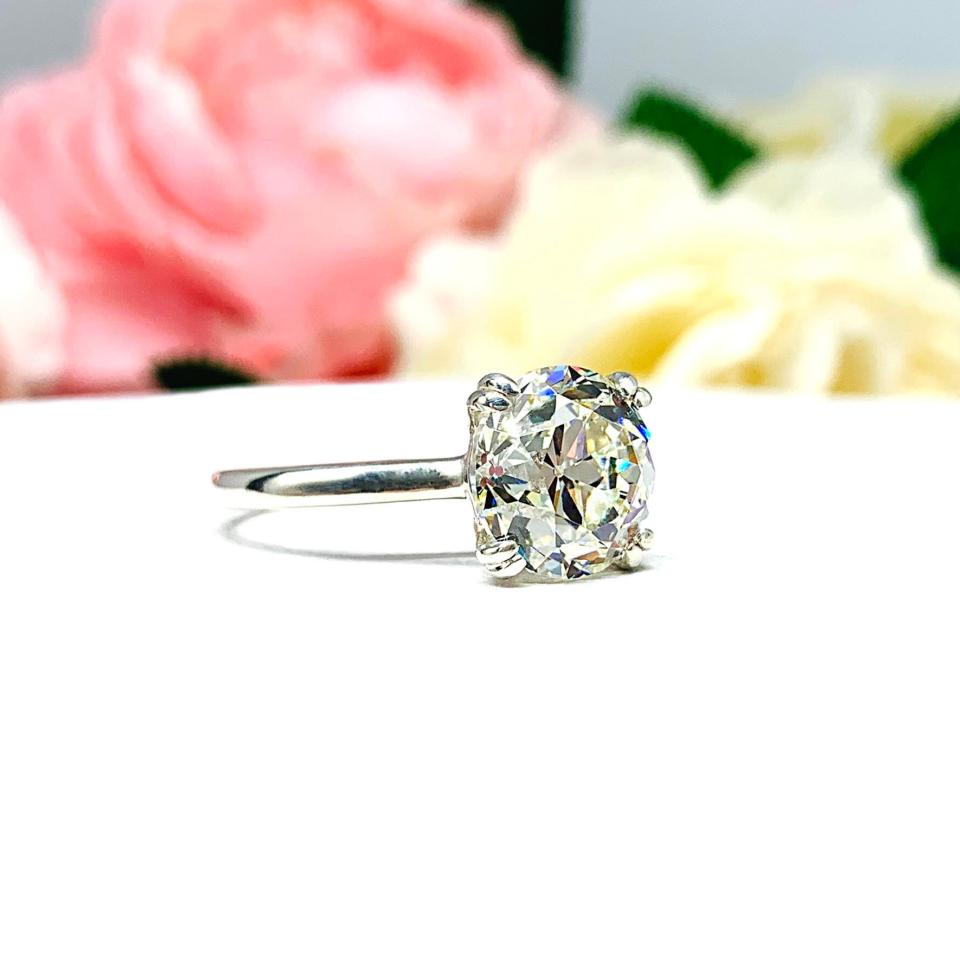 A romantic close-up of a 10mm (4 ct) old European cut near-colorless CZ solitaire engagement ring. Set in a sterling silver split prong cathedral-style mount, the ring gleams with vintage-inspired elegance. Soft pink and white flowers blur in the background, adding a dreamy, bridal ambiance.