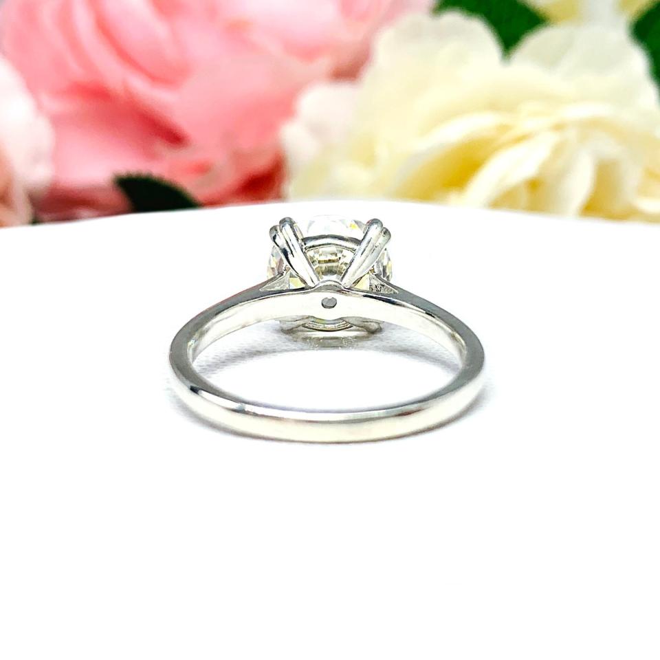 A romantic close-up of a 10mm (4 ct) old European cut near-colorless CZ solitaire engagement ring. Set in a sterling silver split prong cathedral-style mount, the ring gleams with vintage-inspired elegance. Soft pink and white flowers blur in the background, adding a dreamy, bridal ambiance.