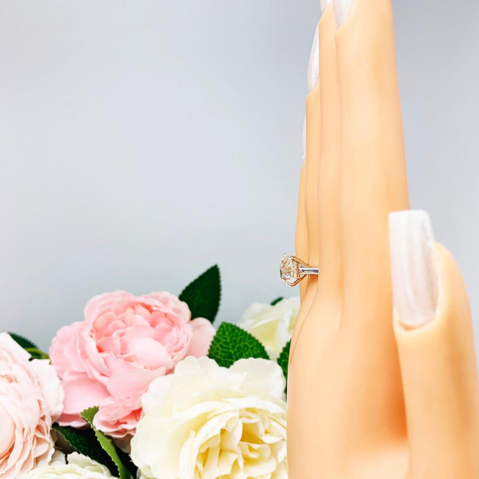 A display hand with long white fingernails standing up and with the top of the hand facing toward the left, displayed against a soft-focus background of pink and white flowers. The display hand is wearing a 10mm (4 ct) old European cut near-colorless CZ solitaire engagement ring. Set in a split prong cathedral-style mount, the ring gleams with vintage-inspired elegance.