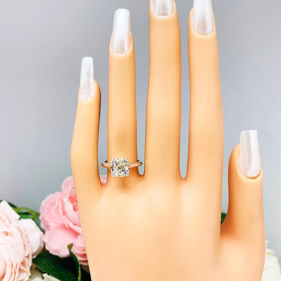 Mannequin hand with long white shimmery nails, displayed against a soft white-gray background with pink and white floral accents. The hand is wearing a sterling silver split-prong cathedral engagement ring set with a near colorless old mine cut cubic zirconia.