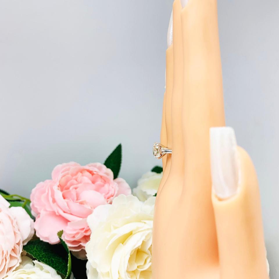 Mannequin hand with long white shimmery nails, displayed against a soft white-gray background with pink and white floral accents. The hand is wearing a sterling silver split-prong cathedral engagement ring set with a near colorless old mine cut cubic zirconia.