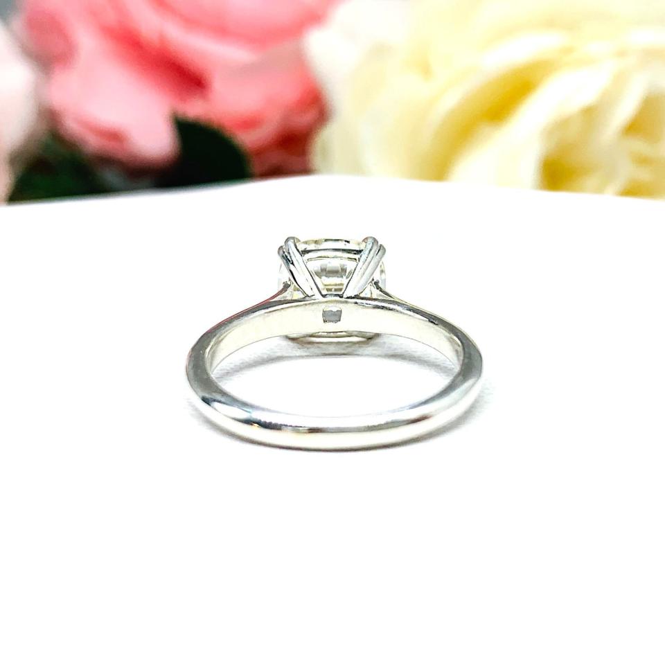 A sterling silver split-prong cathedral engagement ring set with a near colorless old mine cut cubic zirconia. The ring is set against a white background with a blurred image of pink and white flowers in the background.