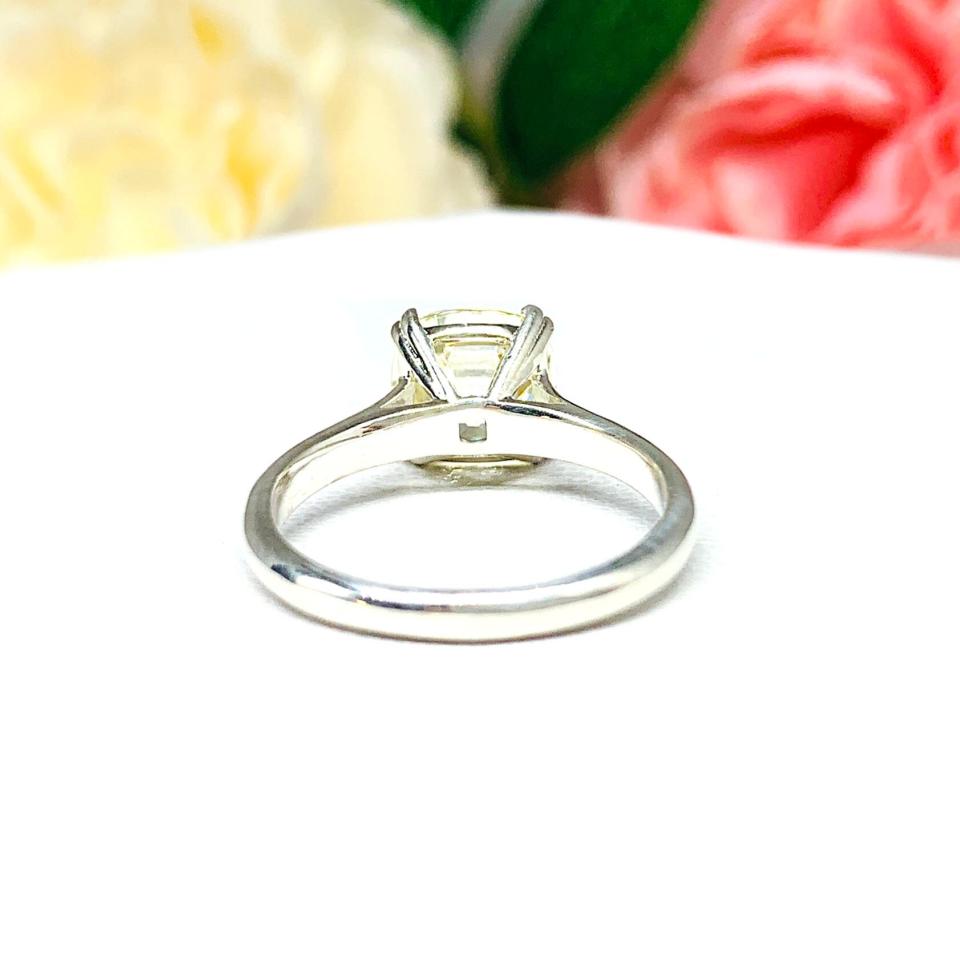 Sterling silver split prong cathedral engagement ring set with an 8mm light yellow old mine cut cubic zirconia. The ring is resting on a white surface with a background of pink and white flowers.