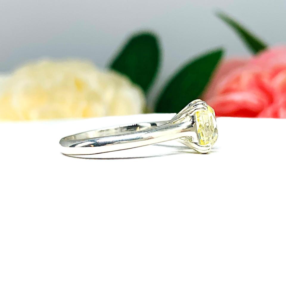Sterling silver split prong cathedral engagement ring set with an 8mm light yellow old mine cut cubic zirconia. The ring is resting on a white surface with a background of pink and white flowers.