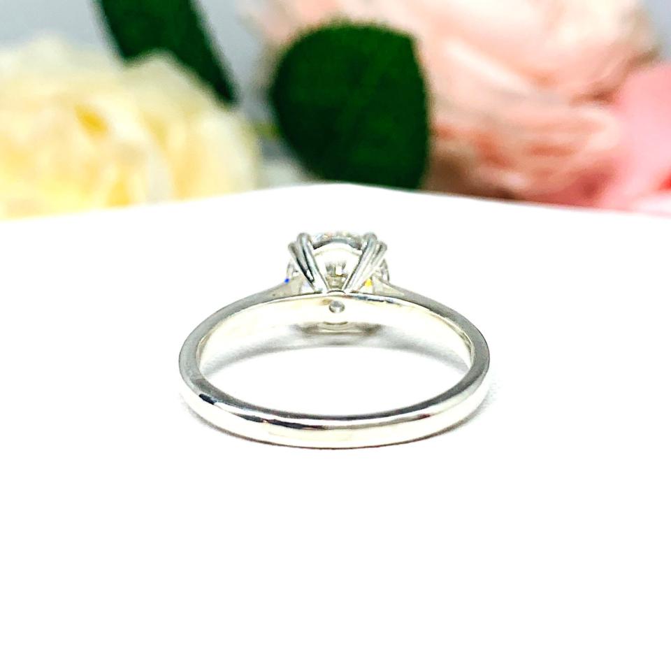 Close-up view of a near colorless (G/H color) round brilliant cut cubic zirconia solitaire ring in a split prong cathedral setting. The ring is made of sterling silver and features a sleek, minimalist design. The setting is simple yet elegant, allowing the center stone to take center stage. The ring is displayed against a soft, blurred background of pastel-colored flowers, creating a romantic and delicate ambiance.