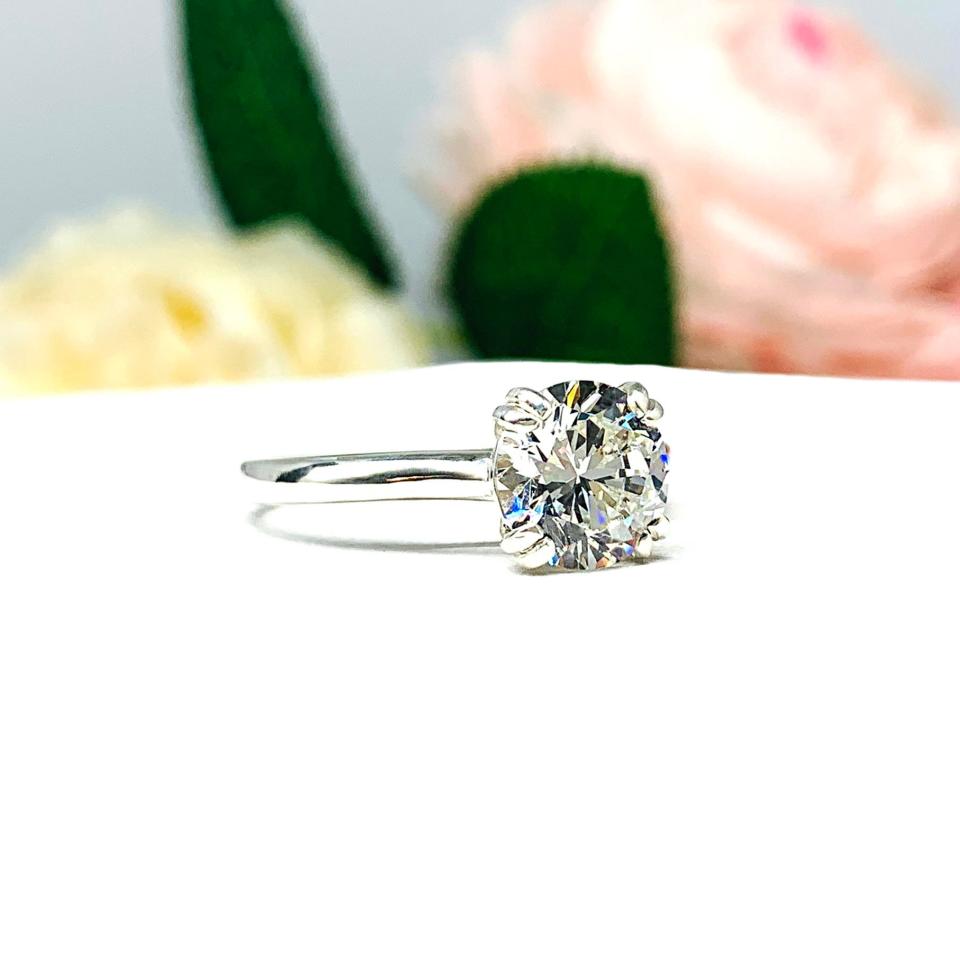 Close-up view of a near colorless (G/H color) round brilliant cut cubic zirconia solitaire ring in a split prong cathedral setting. The ring is made of sterling silver and features a sleek, minimalist design. The setting is simple yet elegant, allowing the center stone to take center stage. The ring is displayed against a soft, blurred background of pastel-colored flowers, creating a romantic and delicate ambiance.