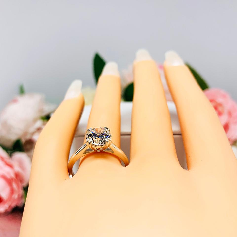 Mannequin hand with long white shimmery nails, displayed against a soft white-gray background with pink and white floral accents. The hand is wearing a near colorless (G/H color) round brilliant cut cubic zirconia solitaire ring in a split prong cathedral setting. The ring is made of sterling silver and features a sleek, minimalist design. The setting is simple yet elegant, allowing the center stone to take center stage.