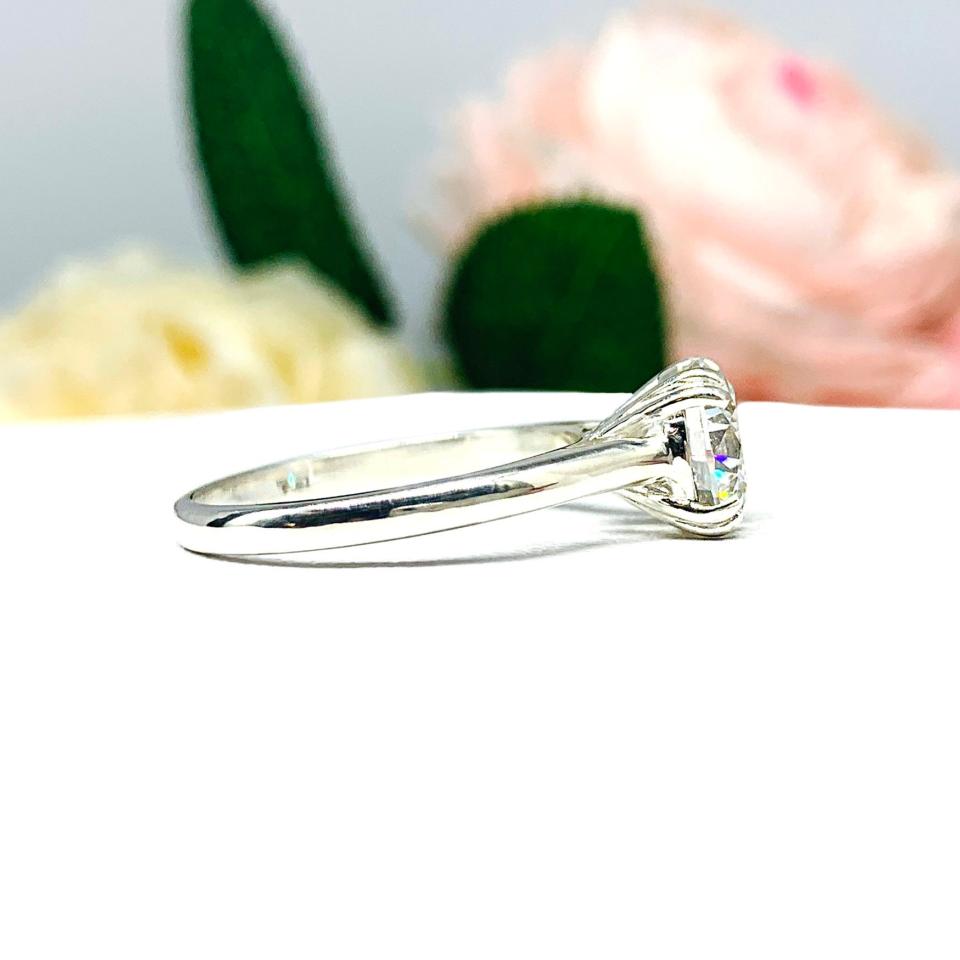 Close-up view of a near colorless (G/H color) round brilliant cut cubic zirconia solitaire ring in a split prong cathedral setting. The ring is made of sterling silver and features a sleek, minimalist design. The setting is simple yet elegant, allowing the center stone to take center stage. The ring is displayed against a soft, blurred background of pastel-colored flowers, creating a romantic and delicate ambiance.