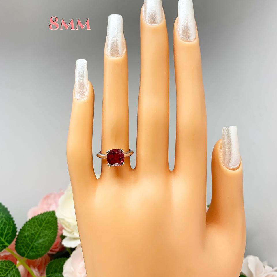 Mannequin hand with long white shimmery nails, displayed against a soft white-gray background with pink and white floral accents. The hand is wearing a sterling silver ring with a cushion cut lab created deep red ruby set in a split-prong setting. The text '8MM' appears above the hand.