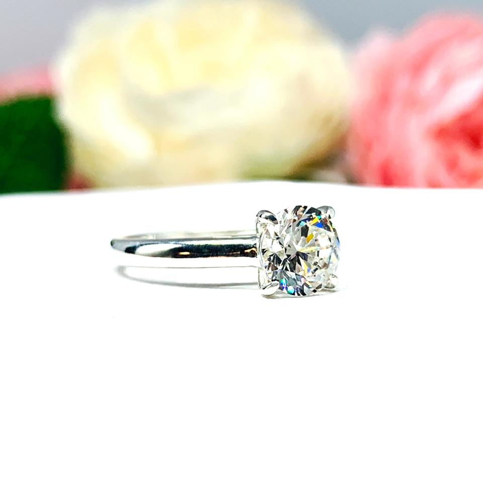 Close-up view of a near colorless (G/H color) round brilliant cut cubic zirconia solitaire ring in a claw prong cathedral setting. The ring is made of sterling silver and features a sleek, minimalist design. The setting is simple yet elegant, allowing the center stone to take center stage. The ring is displayed against a soft, blurred background of pastel-colored flowers, creating a romantic and delicate ambiance.