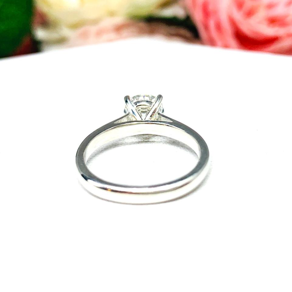 Close-up view of a near colorless (G/H color) round brilliant cut cubic zirconia solitaire ring in a claw prong cathedral setting. The ring is made of sterling silver and features a sleek, minimalist design. The setting is simple yet elegant, allowing the center stone to take center stage. The ring is displayed against a soft, blurred background of pastel-colored flowers, creating a romantic and delicate ambiance.