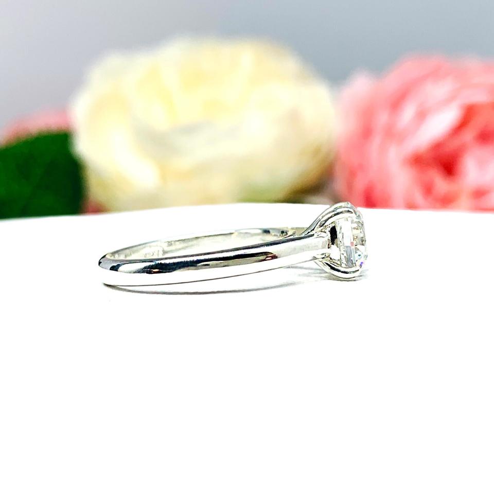 Close-up view of a near colorless (G/H color) round brilliant cut cubic zirconia solitaire ring in a claw prong cathedral setting. The ring is made of sterling silver and features a sleek, minimalist design. The setting is simple yet elegant, allowing the center stone to take center stage. The ring is displayed against a soft, blurred background of pastel-colored flowers, creating a romantic and delicate ambiance.