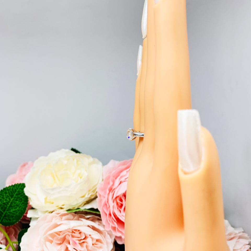 Mannequin hand with long white shimmery nails, displayed against a soft white-gray background with pink and white floral accents. The hand is wearing a near colorless (G/H color) round brilliant cut cubic zirconia solitaire ring in a claw prong cathedral setting. The ring is made of sterling silver and features a sleek, minimalist design. The setting is simple yet elegant, allowing the center stone to take center stage.
