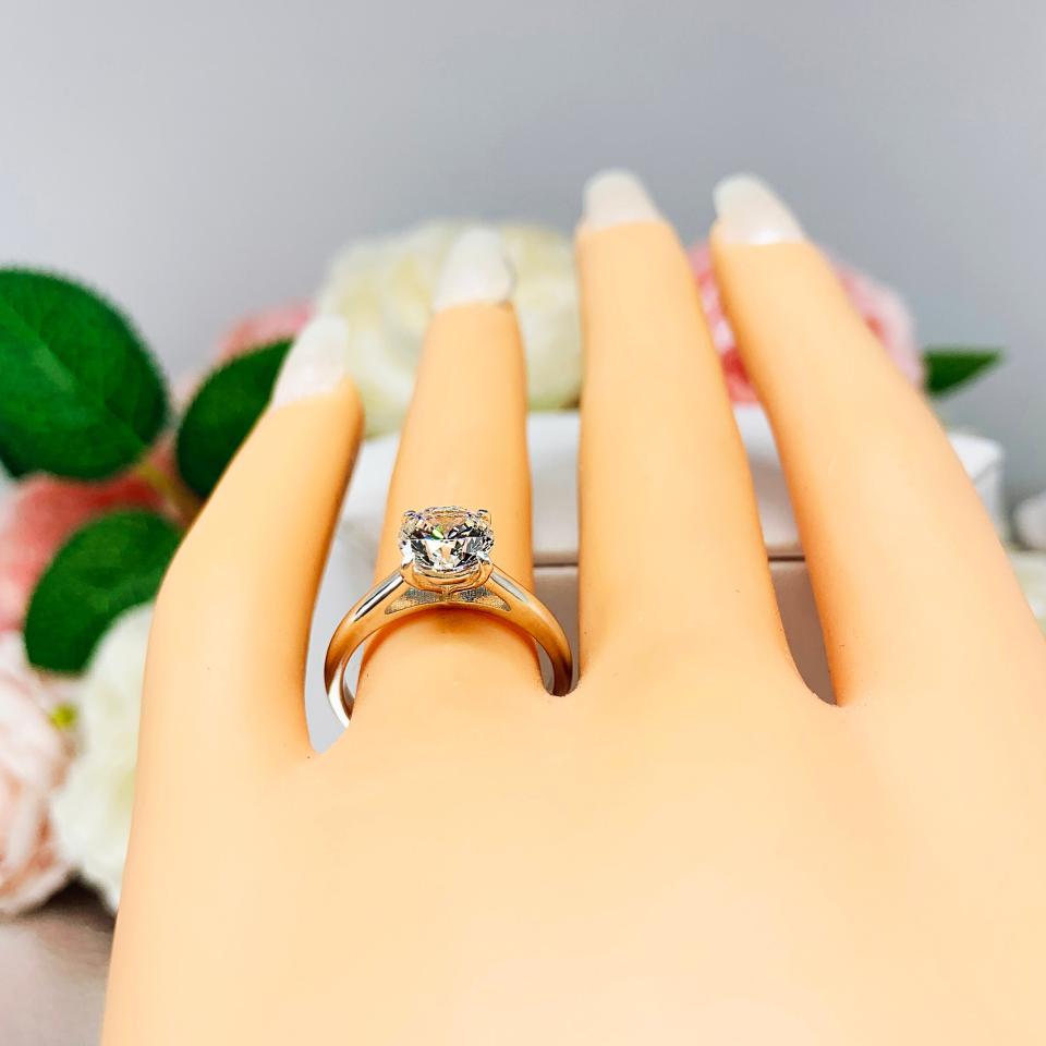 Mannequin hand with long white shimmery nails, displayed against a soft white-gray background with pink and white floral accents. The hand is wearing a near colorless (G/H color) round brilliant cut cubic zirconia solitaire ring in a claw prong cathedral setting. The ring is made of sterling silver and features a sleek, minimalist design. The setting is simple yet elegant, allowing the center stone to take center stage.