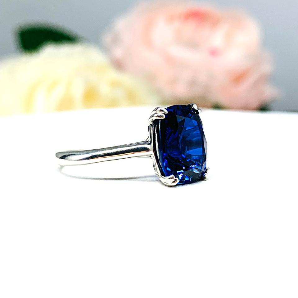 Sterling silver ring featuring a vivid elongated cushion-cut lab created blue sapphire held in a split-prong setting, with soft-focus pink and white flowers in the background.