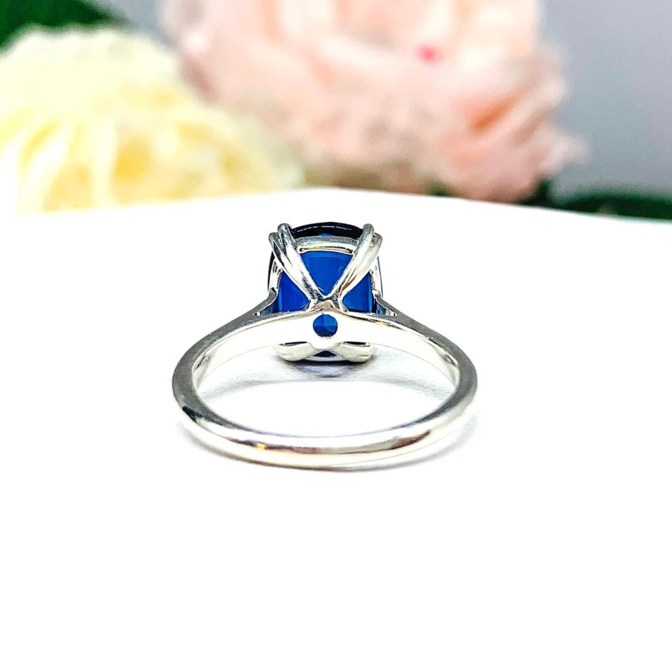 Sterling silver ring featuring a vivid elongated cushion-cut lab created blue sapphire held in a split-prong setting, with soft-focus pink and white flowers in the background.