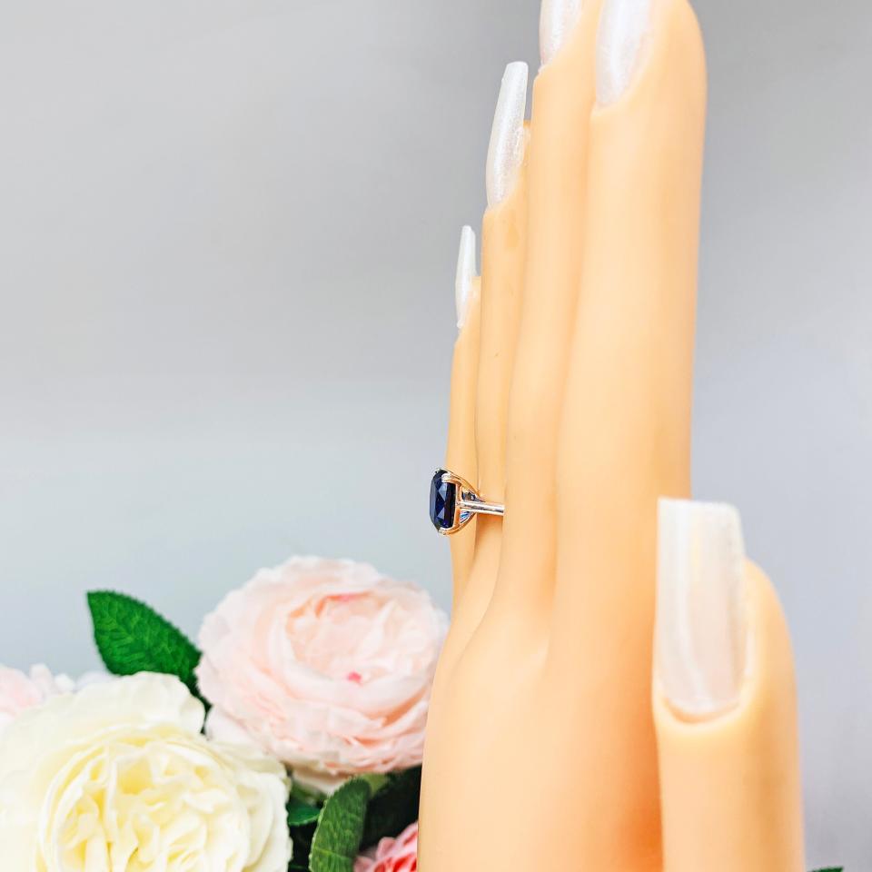 Mannequin hand with long white shimmery nails, displayed against a soft white-gray background with pink and white floral accents. The hand is wearing a sterling silver ring featuring a vivid elongated cushion-cut lab created blue sapphire held in a split-prong setting.