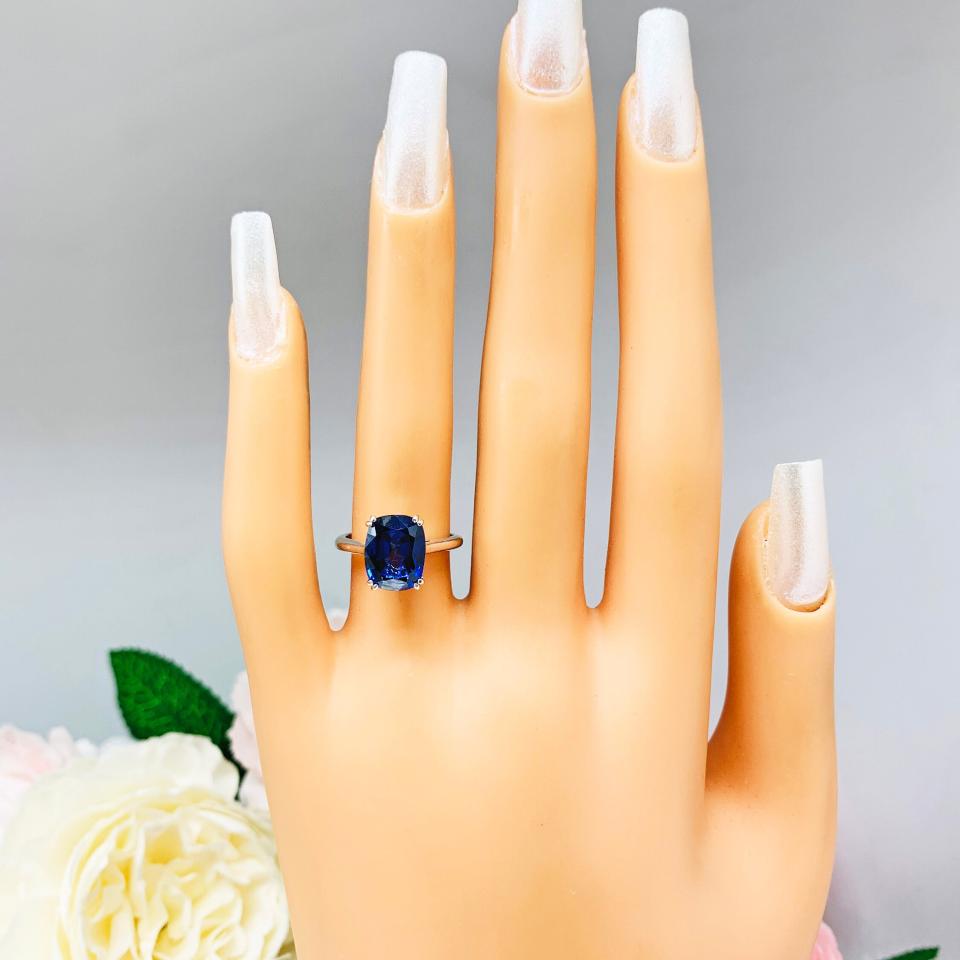 Mannequin hand with long white shimmery nails, displayed against a soft white-gray background with pink and white floral accents. The hand is wearing a sterling silver ring featuring a vivid elongated cushion-cut lab created blue sapphire held in a split-prong setting.
