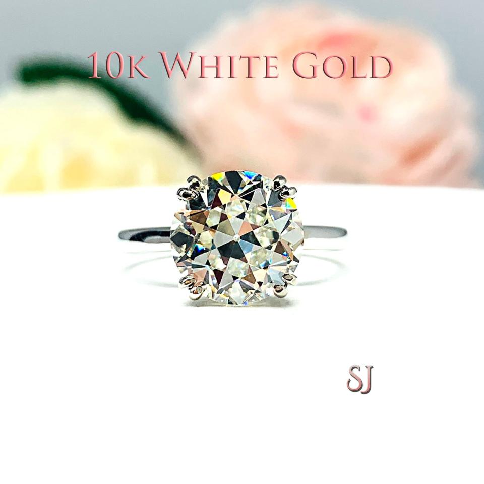 romantic close-up of a 10mm (4 ct) old European cut near-colorless CZ solitaire engagement ring. Set in a 10k white gold split prong cathedral-style mount, the ring gleams with vintage-inspired elegance. Soft pink and white flowers blur in the background, adding a dreamy, bridal ambiance. The text '10K WHITE GOLD' is visible above the ring. The text 'SJ' is visible below the ring.