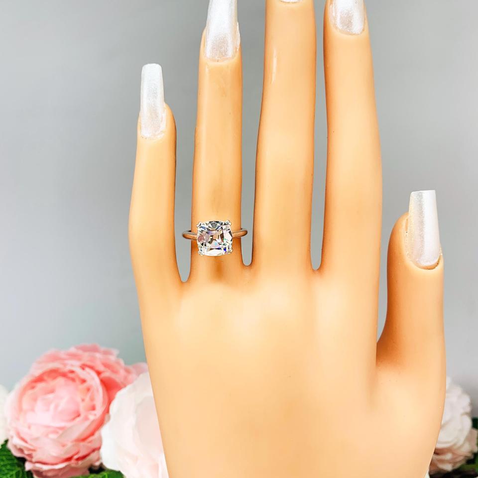 Mannequin hand with long white shimmery nails, displayed against a soft white-gray background with pink and white floral accents. The hand is wearing a sterling silver split-prong cathedral engagement ring set with a near colorless old mine cut cubic zirconia. The text '9MM' appears in the image.