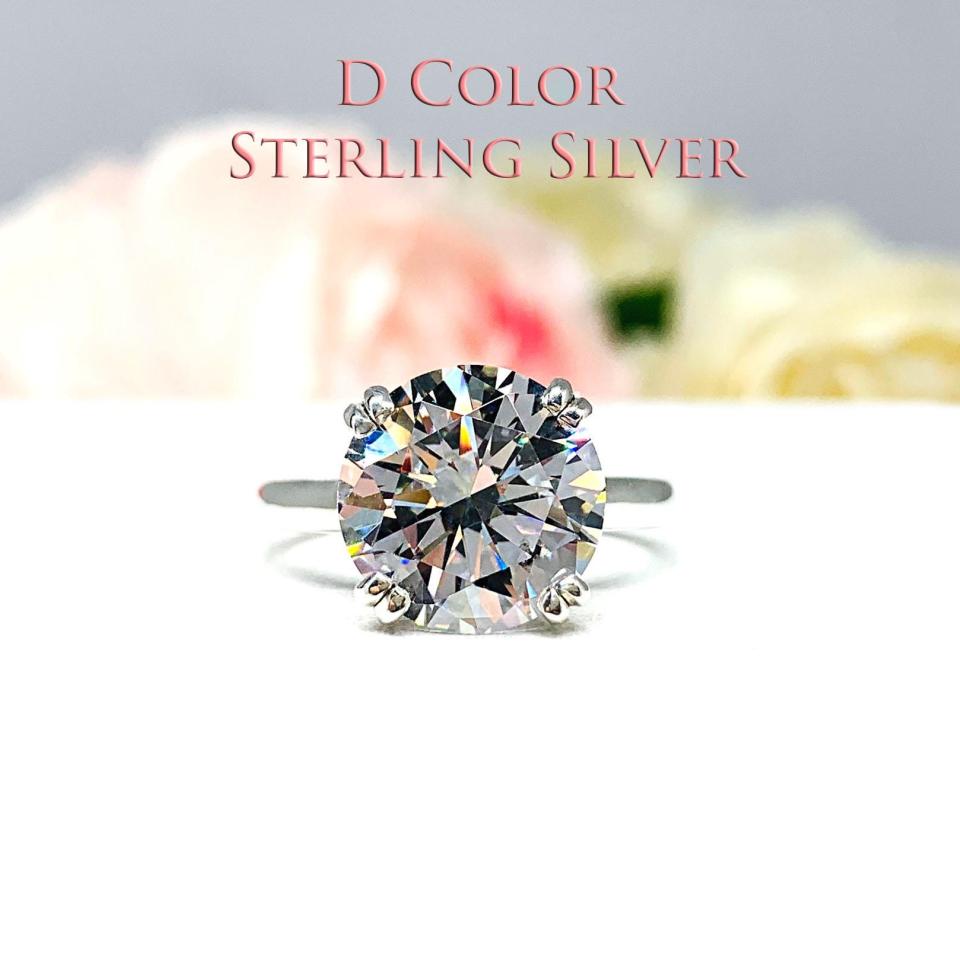 Close-up view of a colorless (D color) round brilliant cut cubic zirconia solitaire ring in a split prong cathedral setting. The ring is made of sterling silver and features a sleek, minimalist design. The setting is simple yet elegant, allowing the center stone to take center stage. The ring is displayed against a soft, blurred background of pastel-colored flowers, creating a romantic and delicate ambiance.