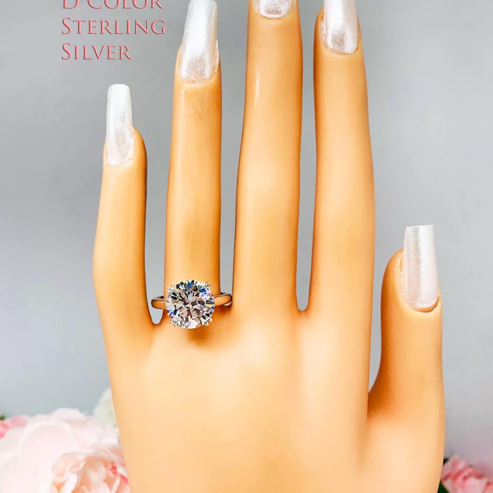 Mannequin hand with long white shimmery nails, displayed against a soft white-gray background with pink and white floral accents. The hand is wearing a colorless (D color) round brilliant cut cubic zirconia solitaire ring in a split prong cathedral setting. The ring is made of sterling silver and features a sleek, minimalist design. The setting is simple yet elegant, allowing the center stone to take center stage.