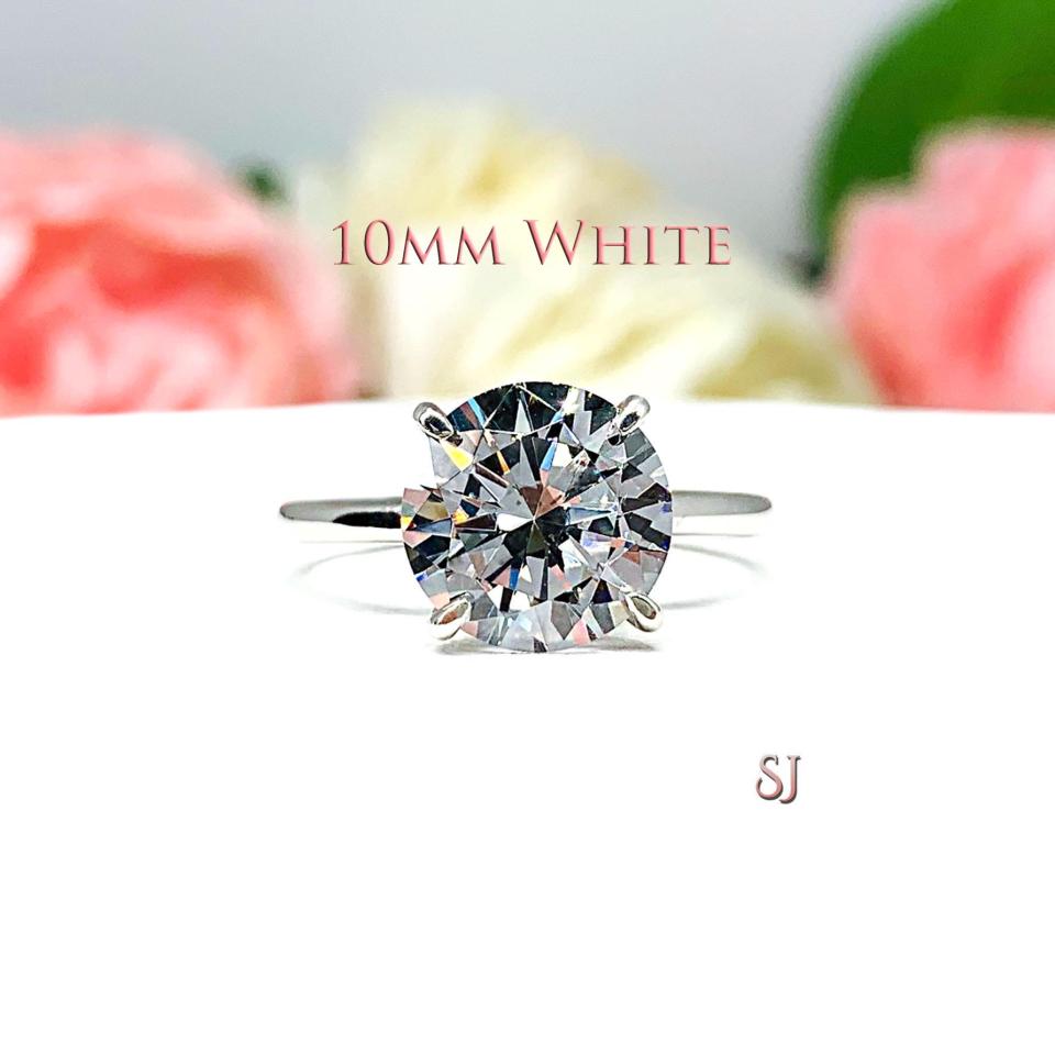 Close-up view of a near colorless (G/H color) round brilliant cut cubic zirconia solitaire ring in a claw prong cathedral setting. The ring is made of sterling silver and features a sleek, minimalist design. The setting is simple yet elegant, allowing the center stone to take center stage. The ring is displayed against a soft, blurred background of pastel-colored flowers, creating a romantic and delicate ambiance. This example is the 10mm white version.