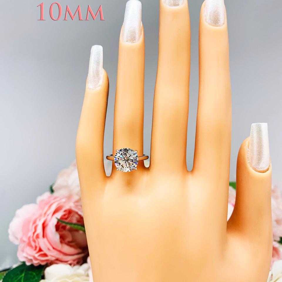 Mannequin hand with long white shimmery nails, displayed against a soft white-gray background with pink and white floral accents. The hand is wearing a near colorless (G/H color) round brilliant cut cubic zirconia solitaire ring in a claw prong cathedral setting. The ring is made of sterling silver and features a sleek, minimalist design. The setting is simple yet elegant, allowing the center stone to take center stage. This example is the 10mm white version.
