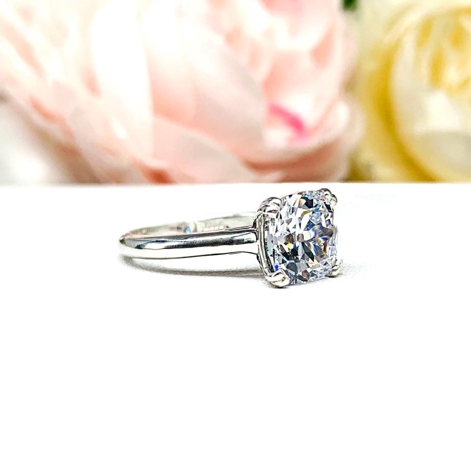 A sterling silver split-prong cathedral engagement ring set with a colorless old mine cut cubic zirconia. The ring is set against a white background with a blurred image of pink and white flowers in the background.