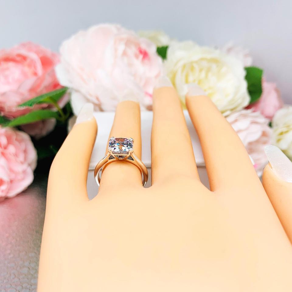 Mannequin hand with long white shimmery nails, displayed against a soft white-gray background with pink and white floral accents. The hand is wearing a sterling silver split-prong cathedral engagement ring set with a colorless old mine cut cubic zirconia.