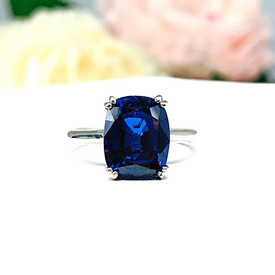 Sterling silver ring featuring a vivid elongated cushion-cut lab created blue sapphire held in a split-prong setting, with soft-focus pink and white flowers in the background.