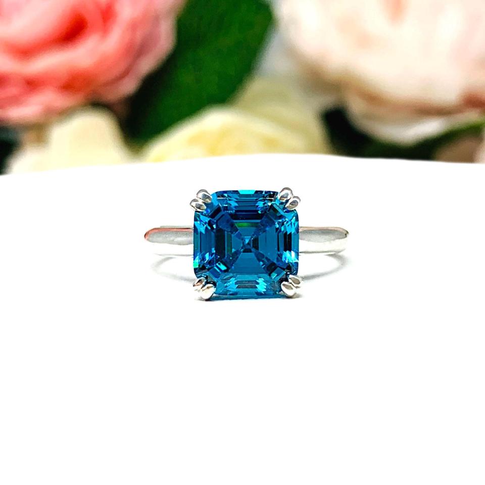 Sterling silver 8mm teal blue asscher cut cubic zirconia solitaire ring. The ring is resting on a white surface with a background of pink and white flowers.