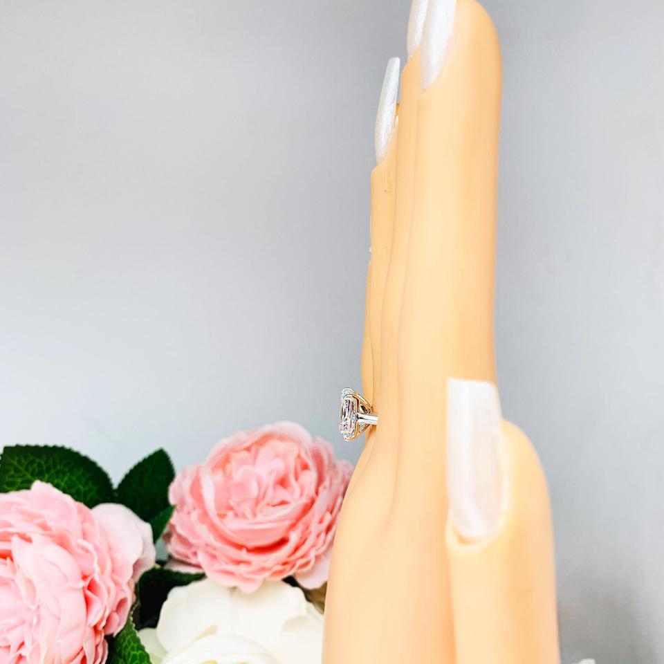 Mannequin hand with long white shimmery nails, displayed against a soft white-gray background with pink and white floral accents. The hand is wearing an elegant sterling silver cathedral engagement ring set with an 10x8mm radiant cut cubic zirconia.