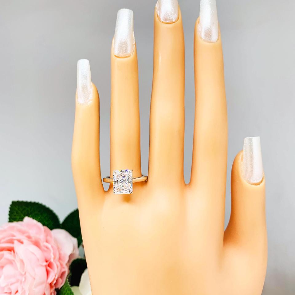 Mannequin hand with long white shimmery nails, displayed against a soft white-gray background with pink and white floral accents. The hand is wearing an elegant sterling silver cathedral engagement ring set with an 10x8mm radiant cut cubic zirconia.