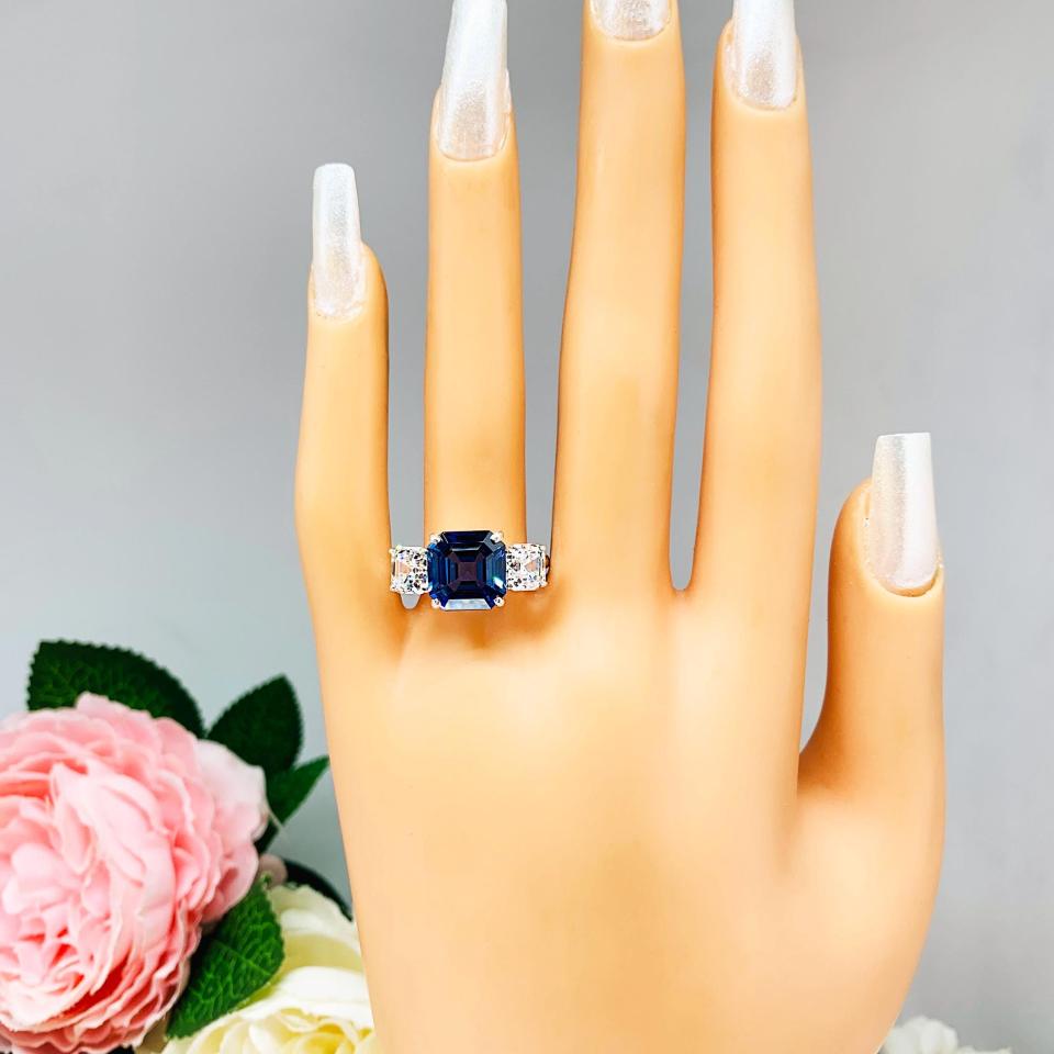 Mannequin hand with long white shimmery nails, displayed against a soft white-gray background with pink and white floral accents. The hand is wearing a sterling silver ring featuring an elegant sterling silver 3-stone ring with a 10mm Asscher‑cut lab blue sapphire center and 6mm CZ side stones.
