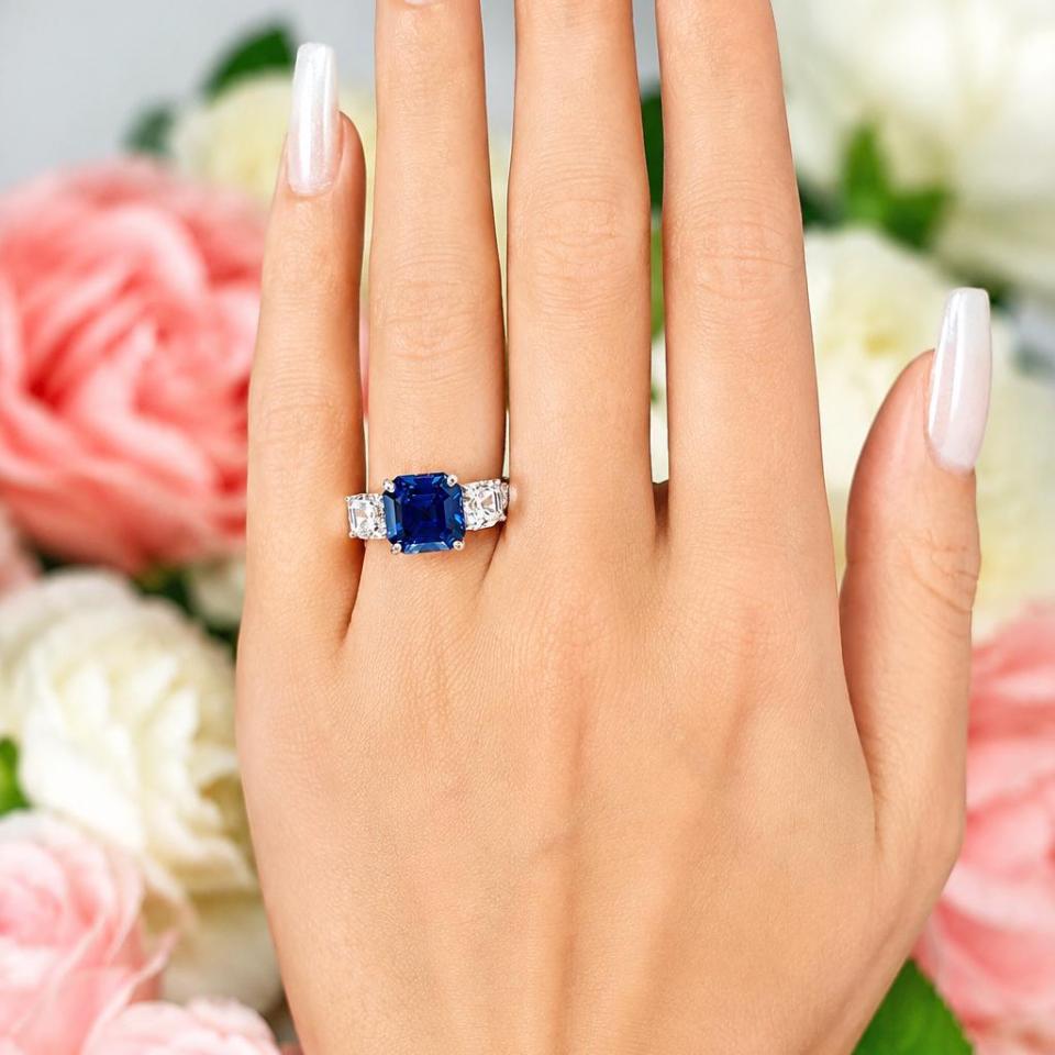 A woman's hand wearing a sterling silver ring featuring an elegant sterling silver 3-stone ring with a 10mm Asscher‑cut lab blue sapphire center and 6mm CZ side stones. Model image for size and styling reference — not a photo of the actual ring.