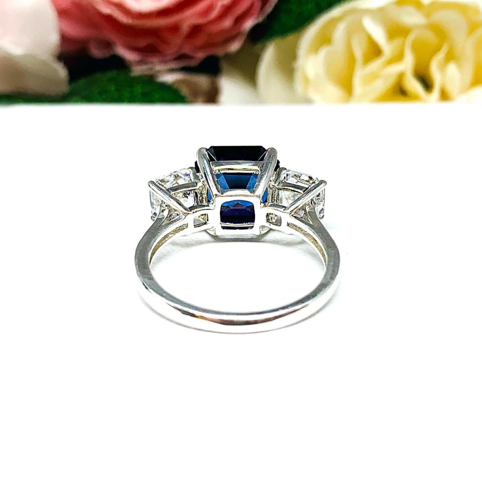 Elegant sterling silver 3-stone ring with a 10mm Asscher‑cut lab blue sapphire center and 6mm CZ side stones, displayed with soft floral backdrop.