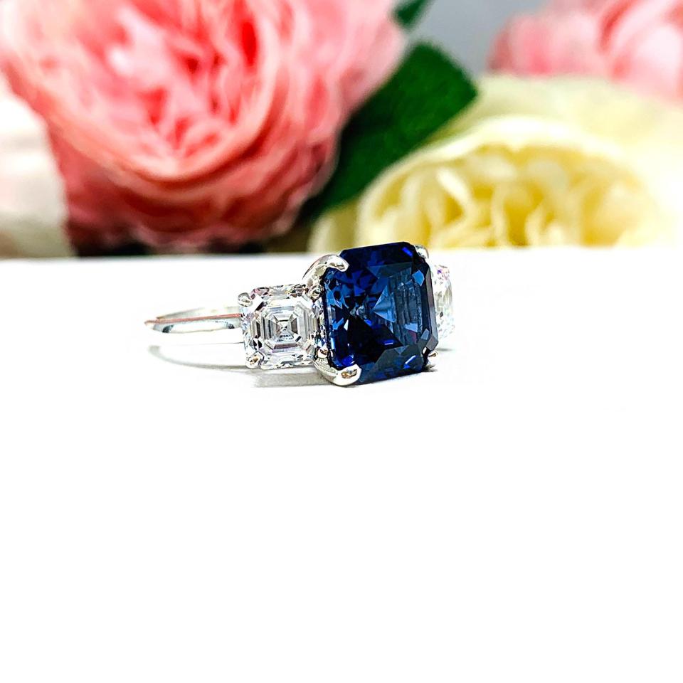 Elegant sterling silver 3-stone ring with a 10mm Asscher‑cut lab blue sapphire center and 6mm CZ side stones, displayed with soft floral backdrop.