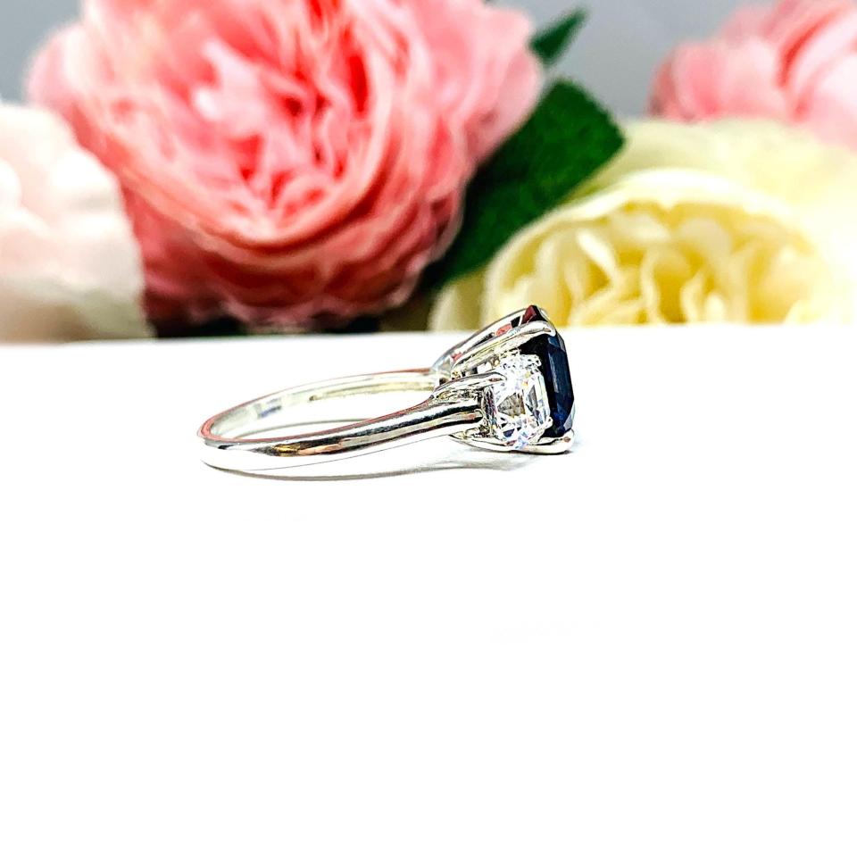 Elegant sterling silver 3-stone ring with a 10mm Asscher‑cut lab blue sapphire center and 6mm CZ side stones, displayed with soft floral backdrop.