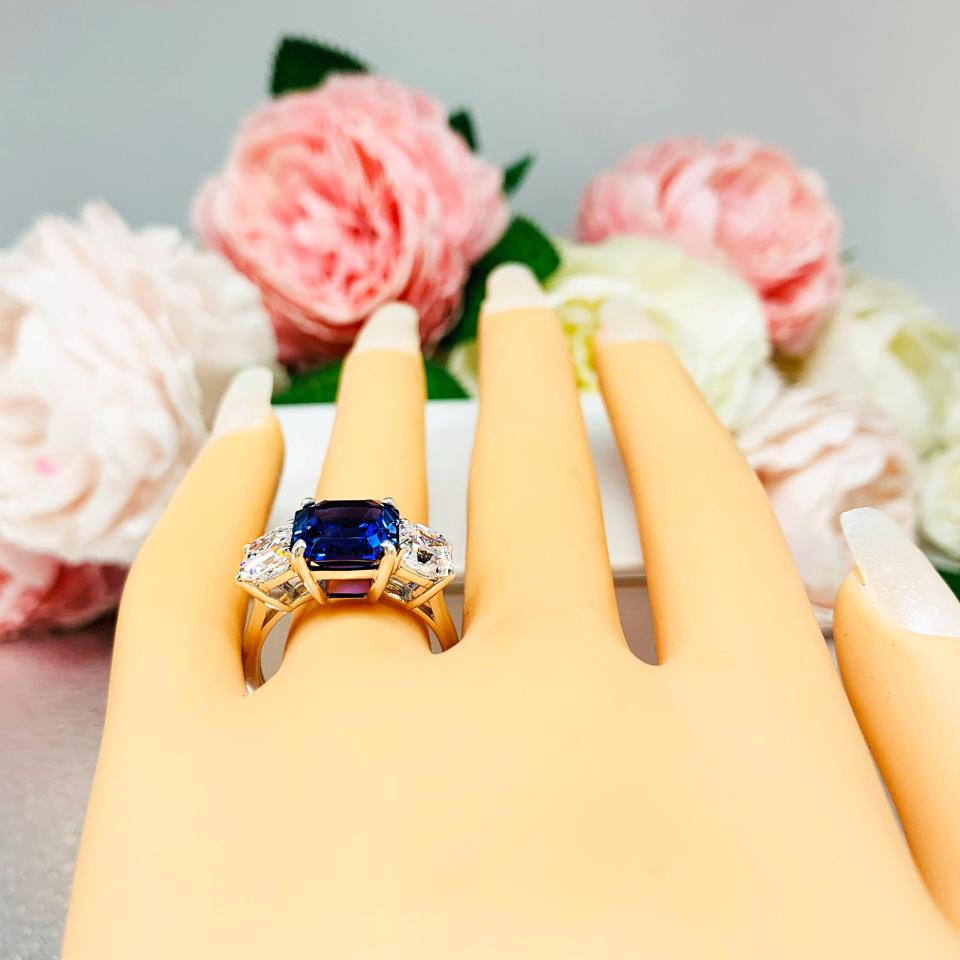 Mannequin hand with long white shimmery nails, displayed against a soft white-gray background with pink and white floral accents. The hand is wearing a sterling silver ring featuring an elegant sterling silver 3-stone ring with a 10mm Asscher‑cut lab blue sapphire center and 6mm CZ side stones.