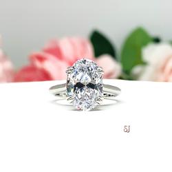 Sterling silver solitaire engagement ring set with a 12x8mm elongated oval cubic zirconia. The ring features split prongs and a cathedral shank. It is resting on a white surface with soft-focus pink and white flowers in the background.