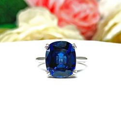 Sterling silver ring featuring a vivid elongated cushion-cut lab created blue sapphire held in a split-prong setting, with soft-focus pink and white flowers in the background.
