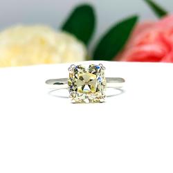 Sterling silver split prong cathedral engagement ring set with an 8mm light yellow old mine cut cubic zirconia. The ring is resting on a white surface with a background of pink and white flowers.